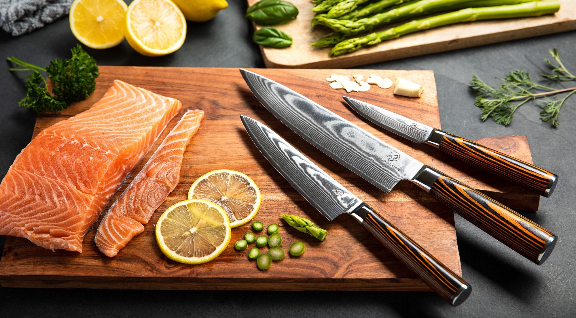 Drei Damastmesser von LH Lifestyle auf einem Brettchen. Ein Stück Lachs liegt links geschnitten. Überhalb des Schneidebretts sind Zitronen und grüner Spargel